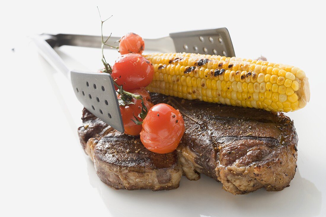 Gegrilltes T-Bone-Steak mit Maiskolben, Tomaten, Grillzange