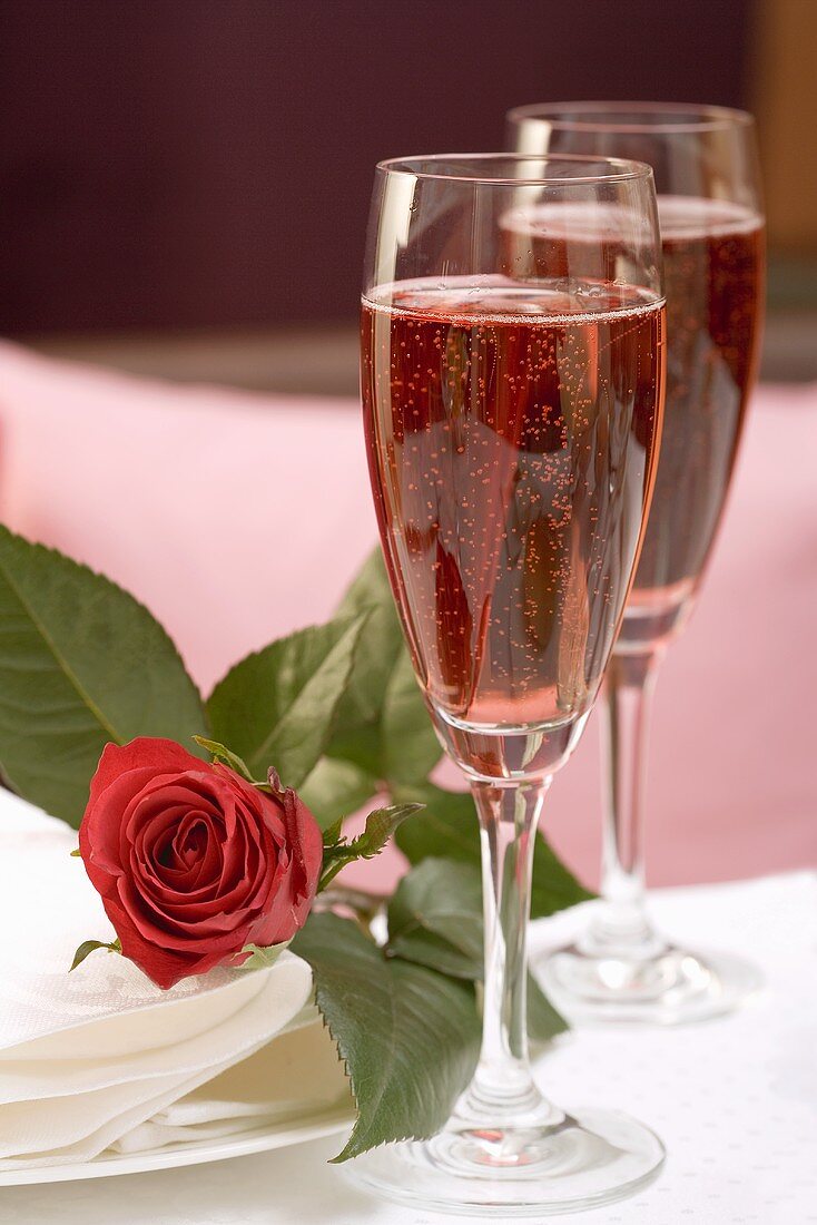 Zwei Gläser rosefarbener Champagner, daneben rote Rose