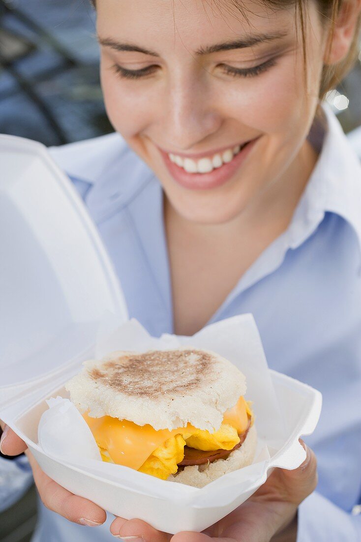 Young woman holding English muffin with cheese, scrambled egg & bacon