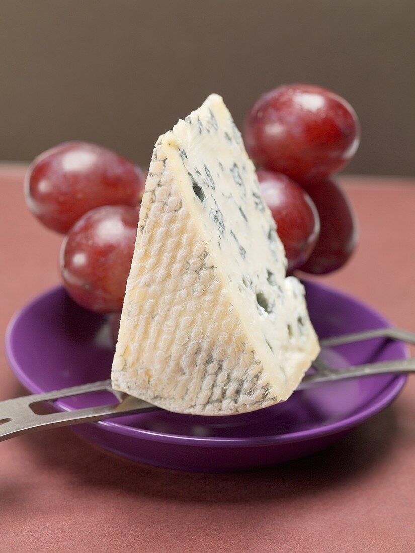 Stück Blauschimmelkäse auf Käsemesser, dahinter rote Trauben