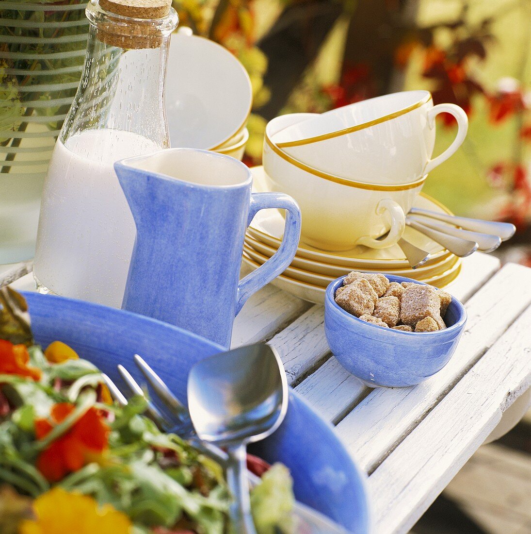 Holztisch mit Salat, Zucker, Milchkanne und Tassen im Freien