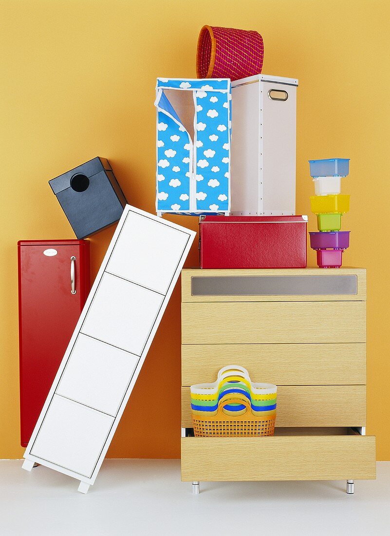 Schubladenkommode, Schränkchen, Schachteln, gestapelte Plastikdosen und Plastikkörbe vor gelber Wand