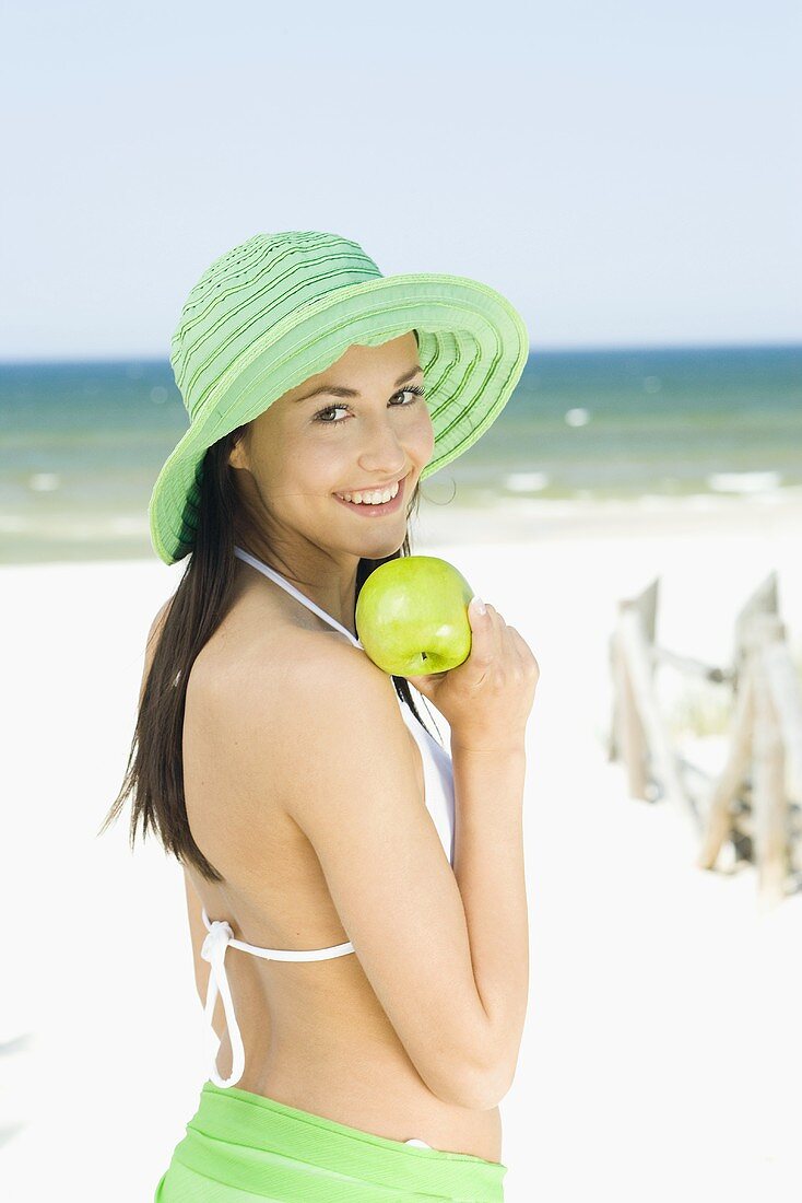 Junge Frau mit Sommerhut und grünem Apfel am Strand