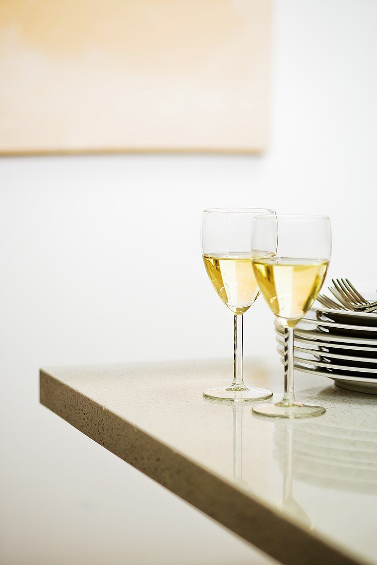 Two glasses of white wine beside pile of plates on table corner