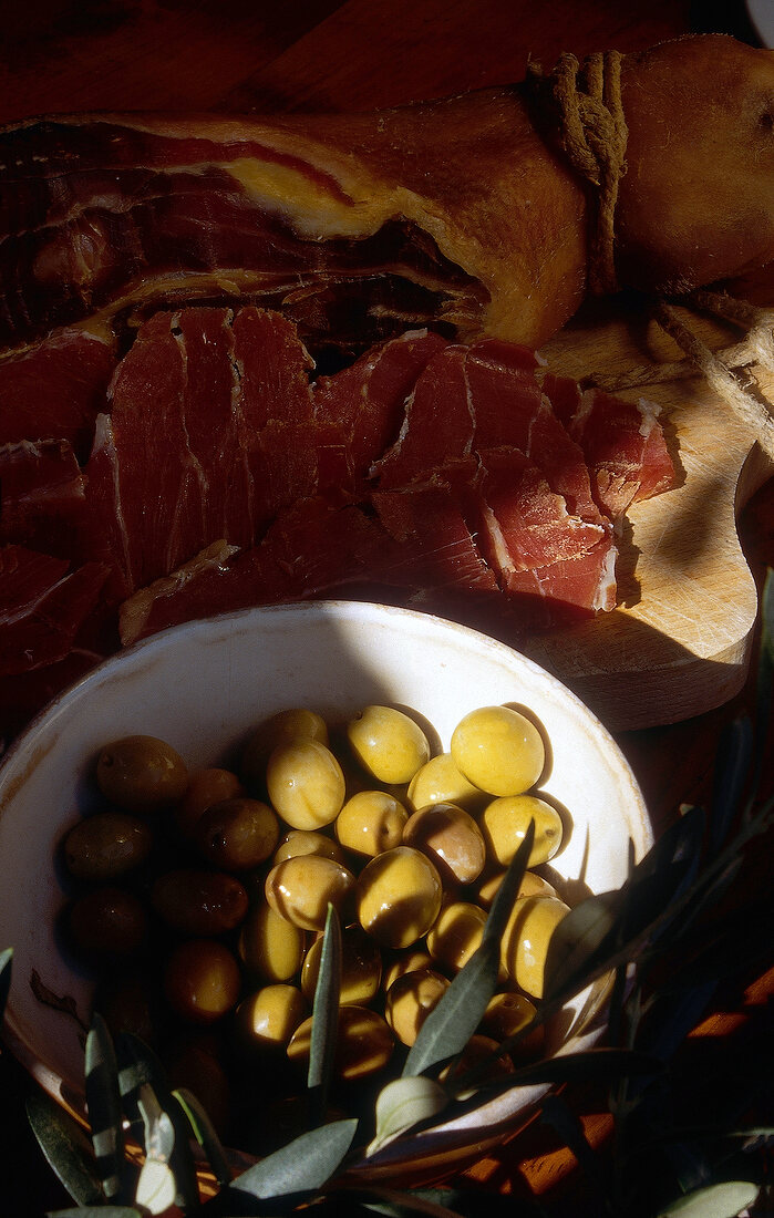 Green olives in bowl with raw ham on chopping board