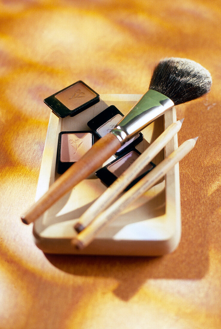 Eye shadow, pencil lip liners and powdered brush kept on wooden bowl