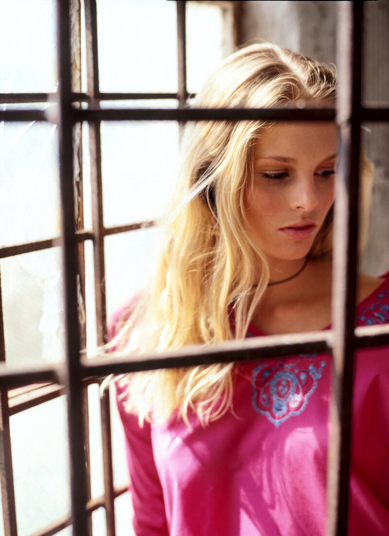 Pensive blonde woman wearing pink top standing at the window, looking downwards