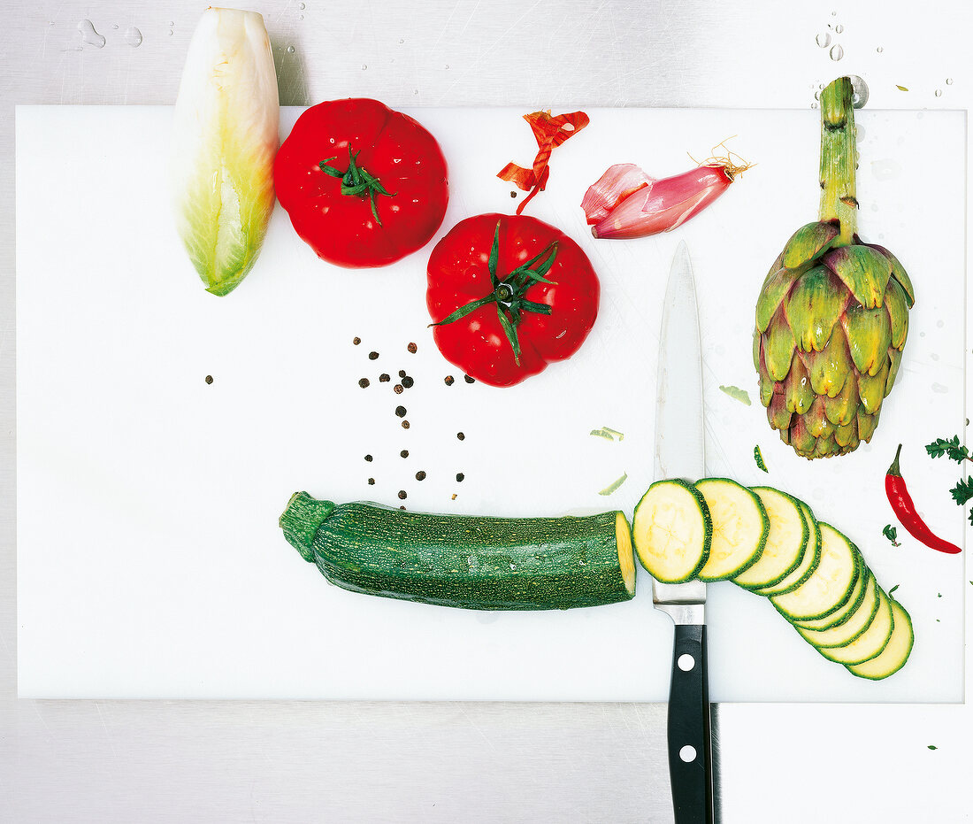 Zucchini in slices with a knife with various vegetables on cutting board, overhead view