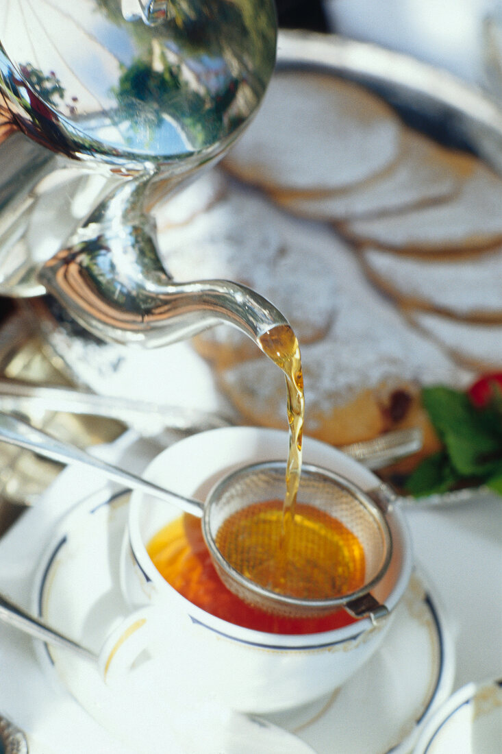 Tea being poured from a silver pot through a sieve into a cup