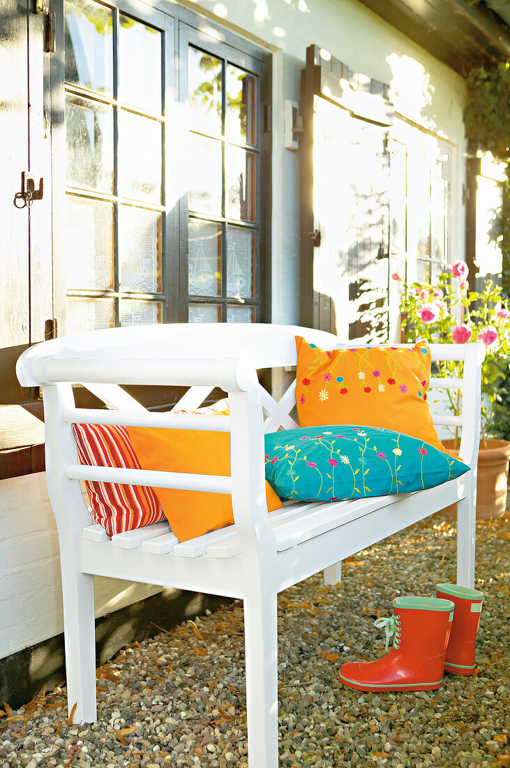 White garden bench on pebbles with colourful cushions and rubber boots on ground