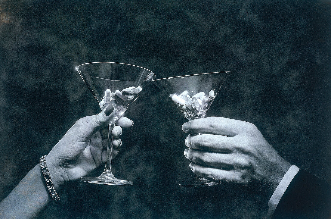 Two hands holding martini glasses with coloured tablets and toasting, black and white
