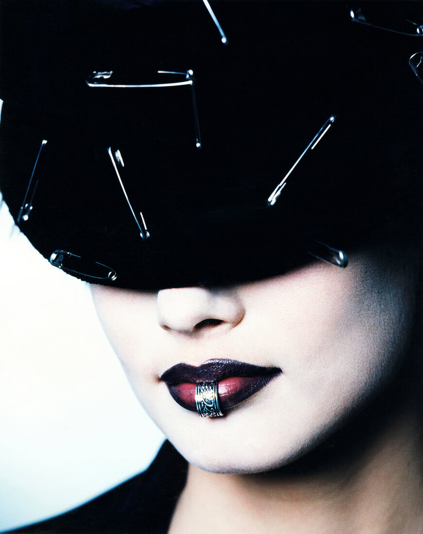 Close-up of glamorous woman with heavy make-up, lip piercing in black hat with safety pins