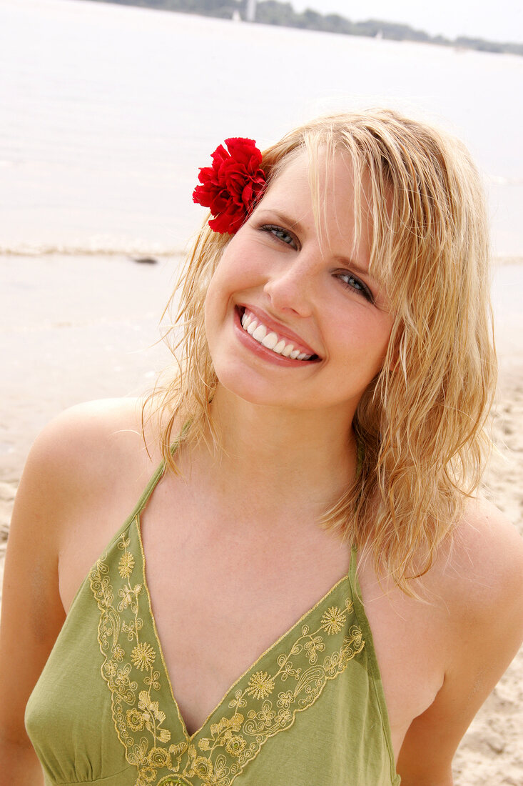 Happy blonde woman with flower in hair wearing green halter top standing on beach, smiling