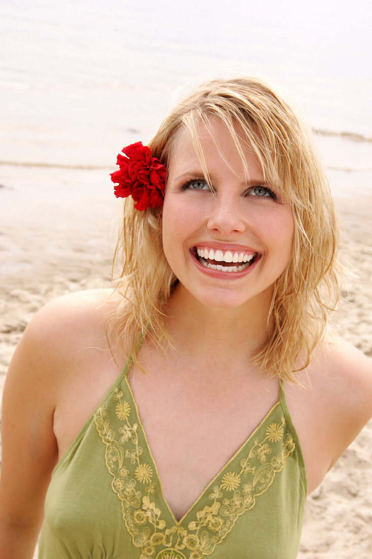 Happy blonde woman with flower in hair wearing green halter top standing on beach, smiling