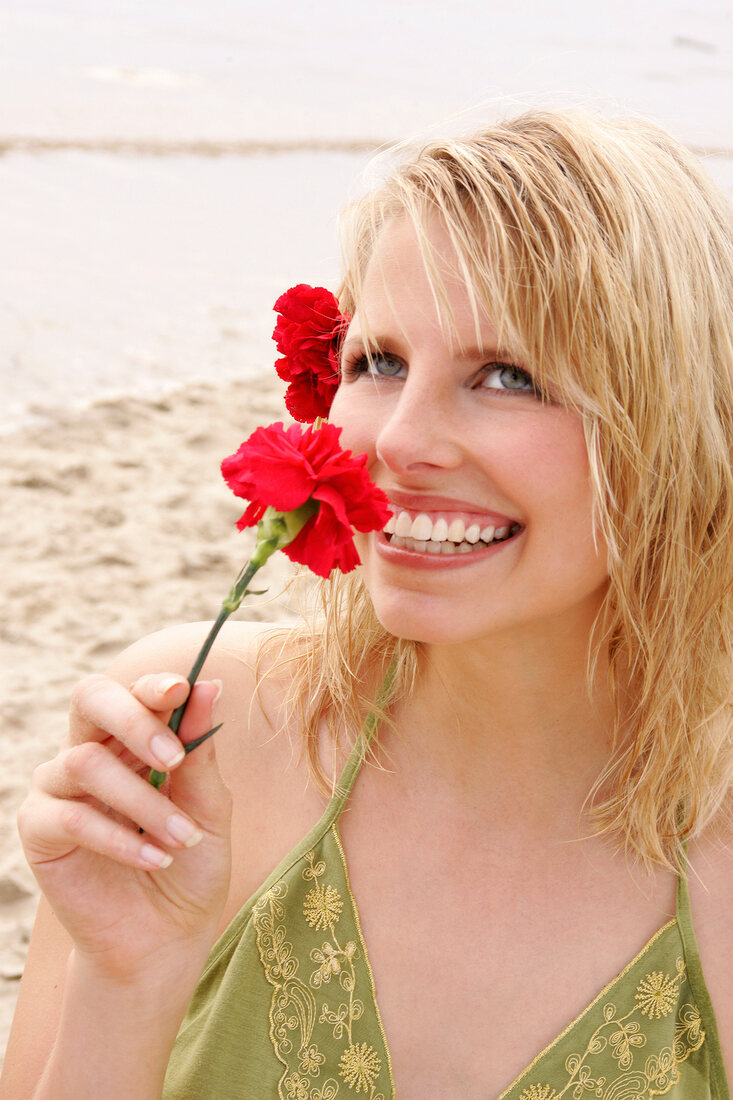 Portrait of beautiful woman wearing green top standing on beach and smelling red carnation