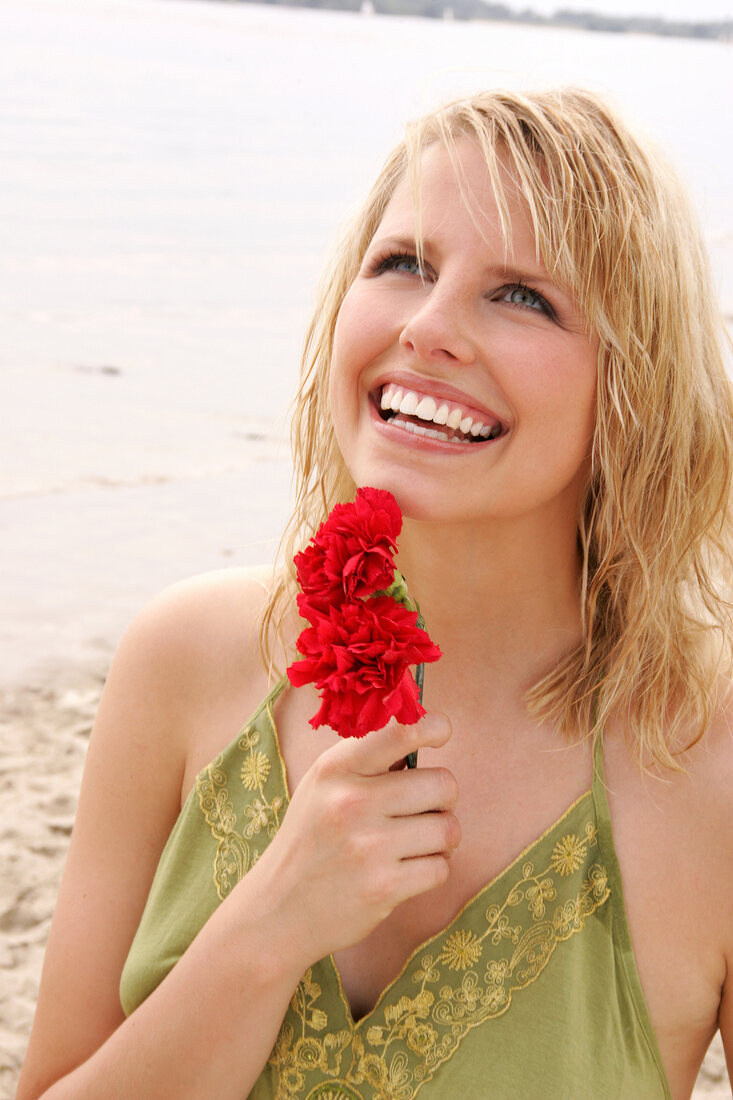 Woman wearing green halterneck dress holding red flower on beach, laughing