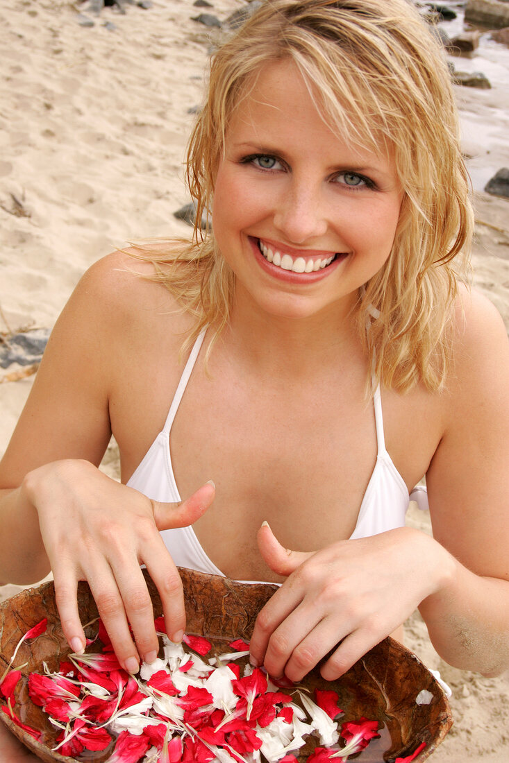 Pretty blonde woman sitting and holding bowl of white and red petals on beach, smiling