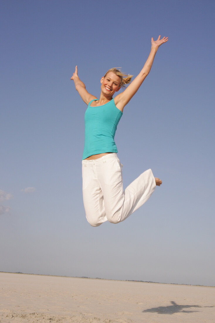 Pretty woman in blue top and white pants jumping on beach with arms outstretched, smiling