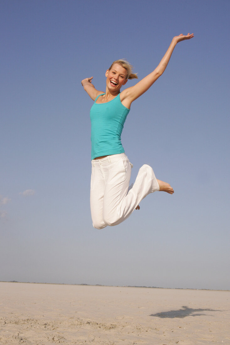 Pretty woman in blue top and white pants jumping on beach with arms outstretched, smiling