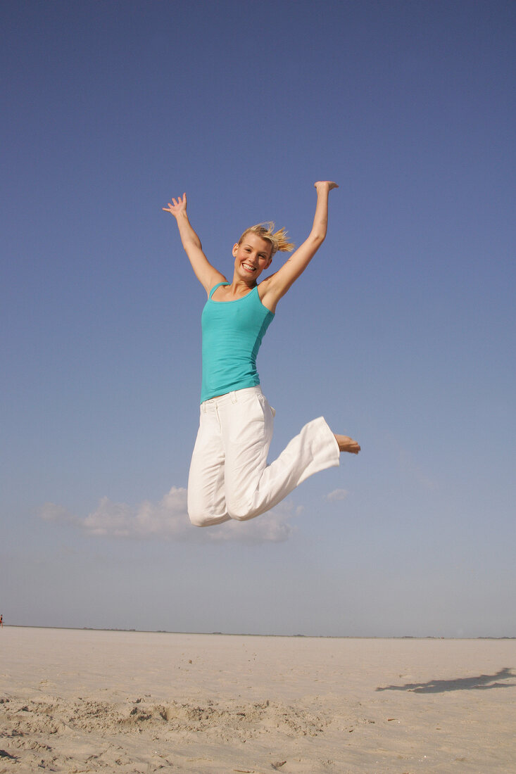 Pretty woman in blue top and white pants jumping on beach with arms outstretched, smiling