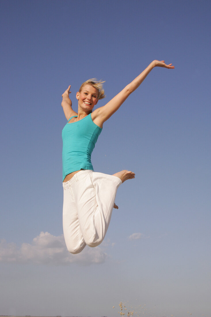 Pretty woman in blue top and white pants jumping on beach with arms outstretched, smiling