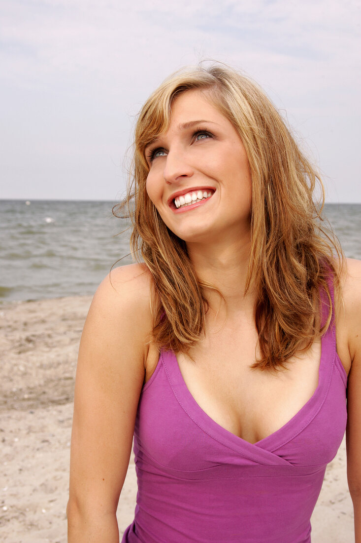 Cheerful blonde woman wearing pink tank top standing on beach, looking up and smiling