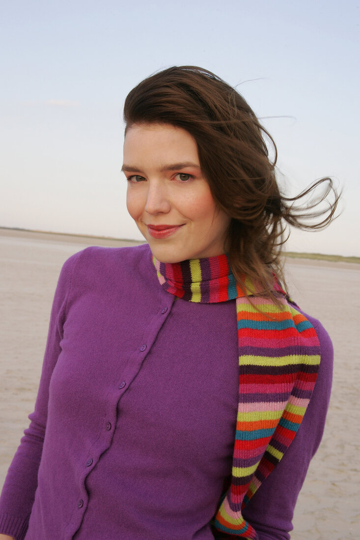 Pretty blonde woman wearing purple sweater and striped scarf standing on beach, smiling
