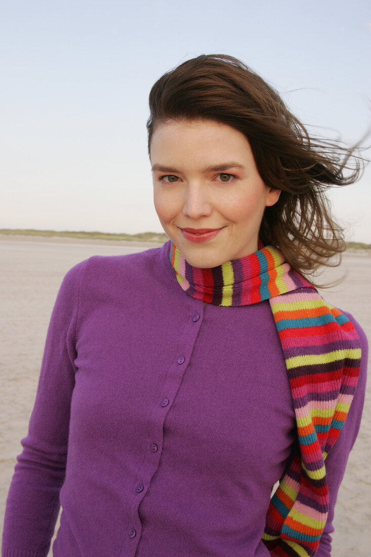 Pretty blonde woman wearing purple sweater and striped scarf standing on beach, smiling