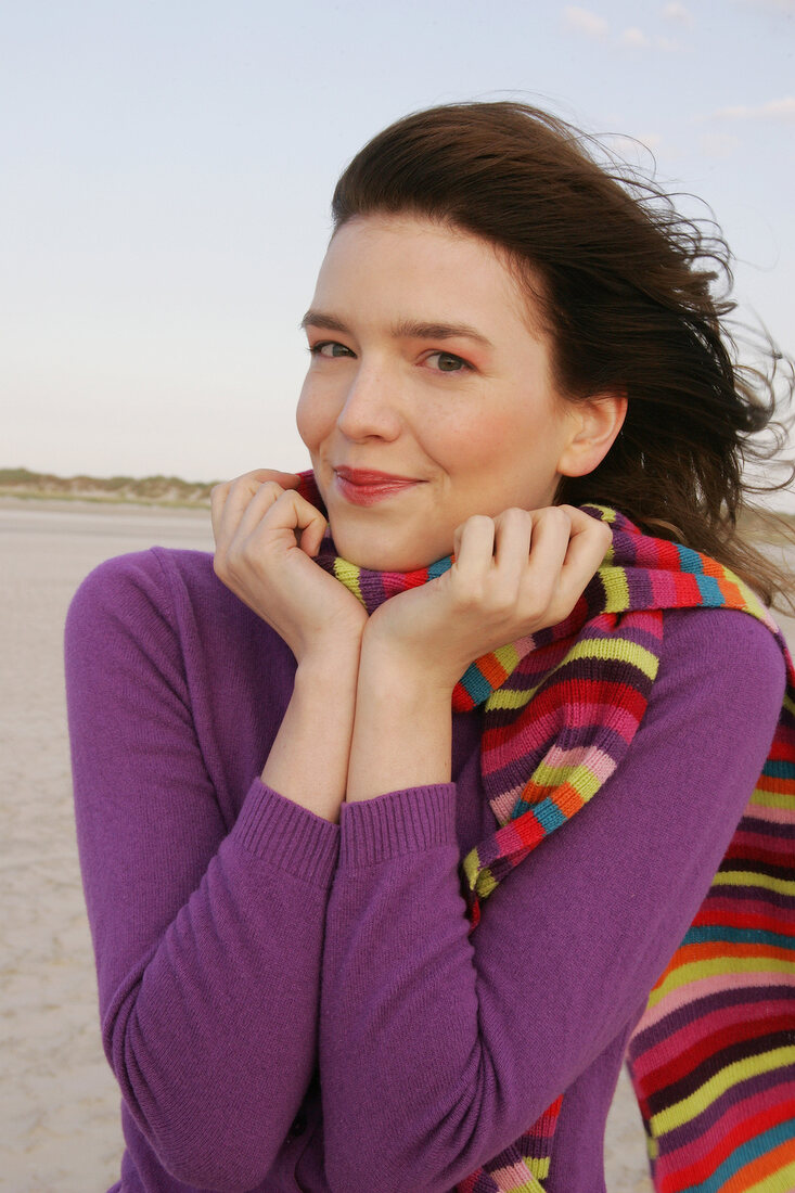 Pretty blonde woman wearing purple sweater and striped scarf standing on beach, smiling