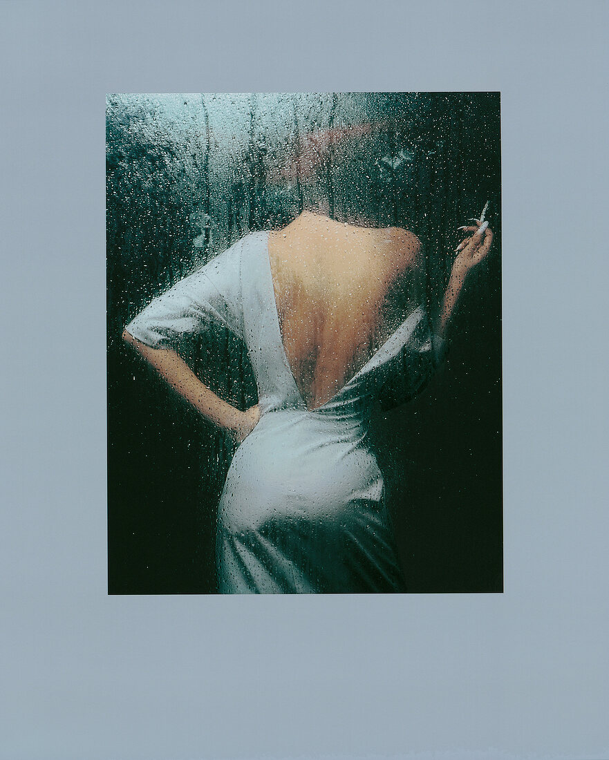 Rear view of glamorous woman in silk taffeta dress standing behind glass with raindrops