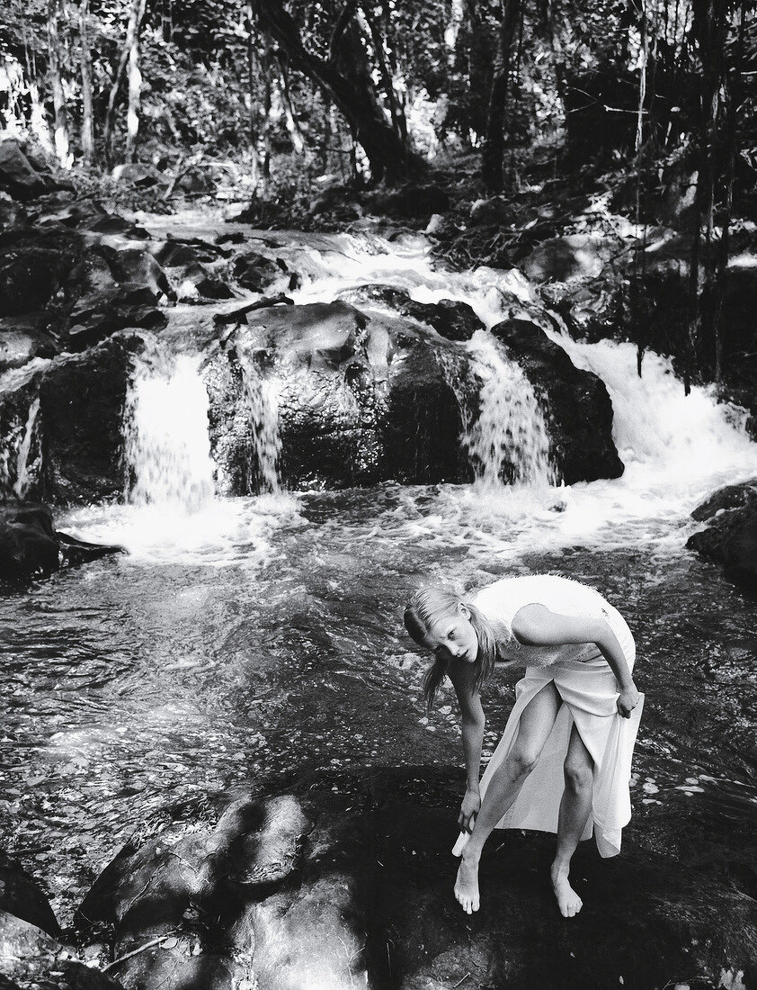 Blonde woman wearing white outfit standing in front of waterfall, black and white