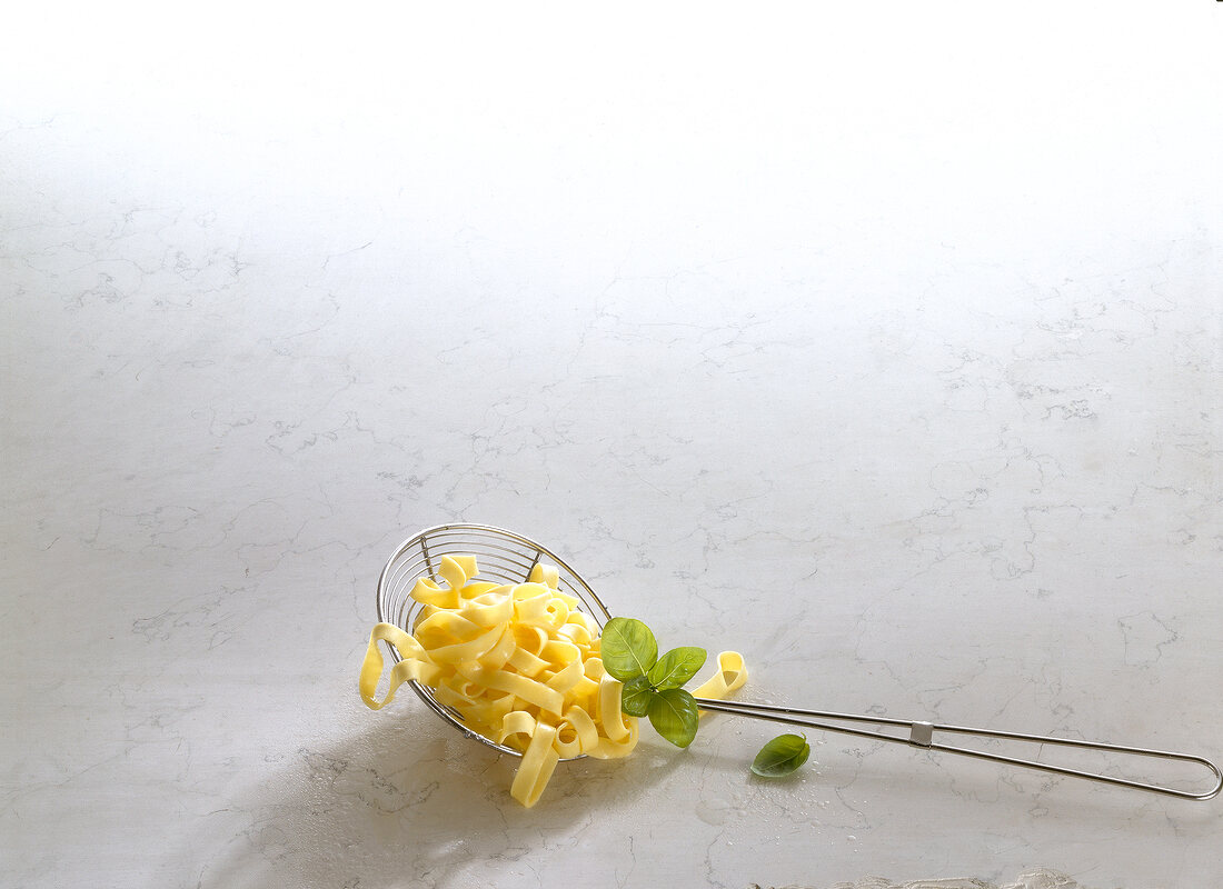 Tagliatelle and basil leaves in strainer on white background