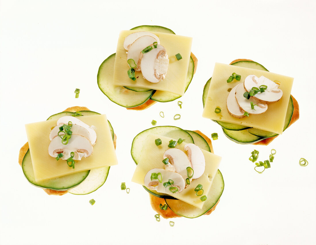 Close-up of four crackers topped with cheese, cucumber slices, mushrooms and chives