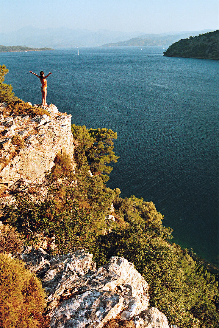 View of woman standing on cliff with arms outstretched at Hamam Bay, Turkey