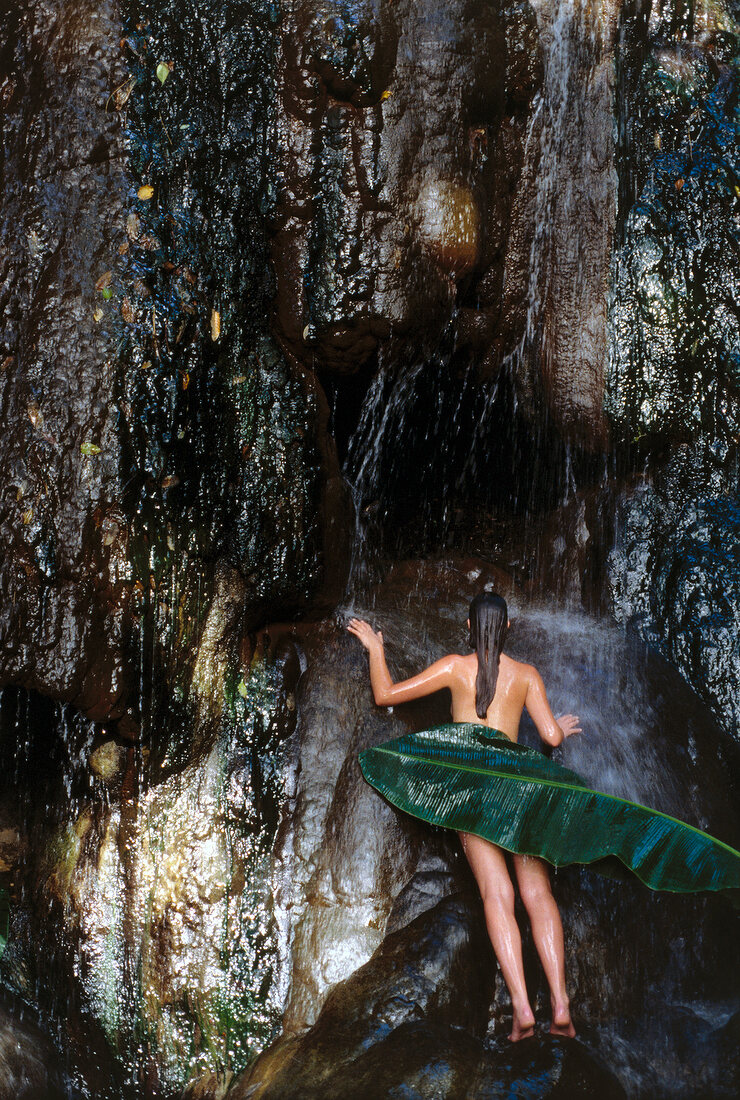 Rear view of nude woman covered with a leaf standing under tropical waterfall