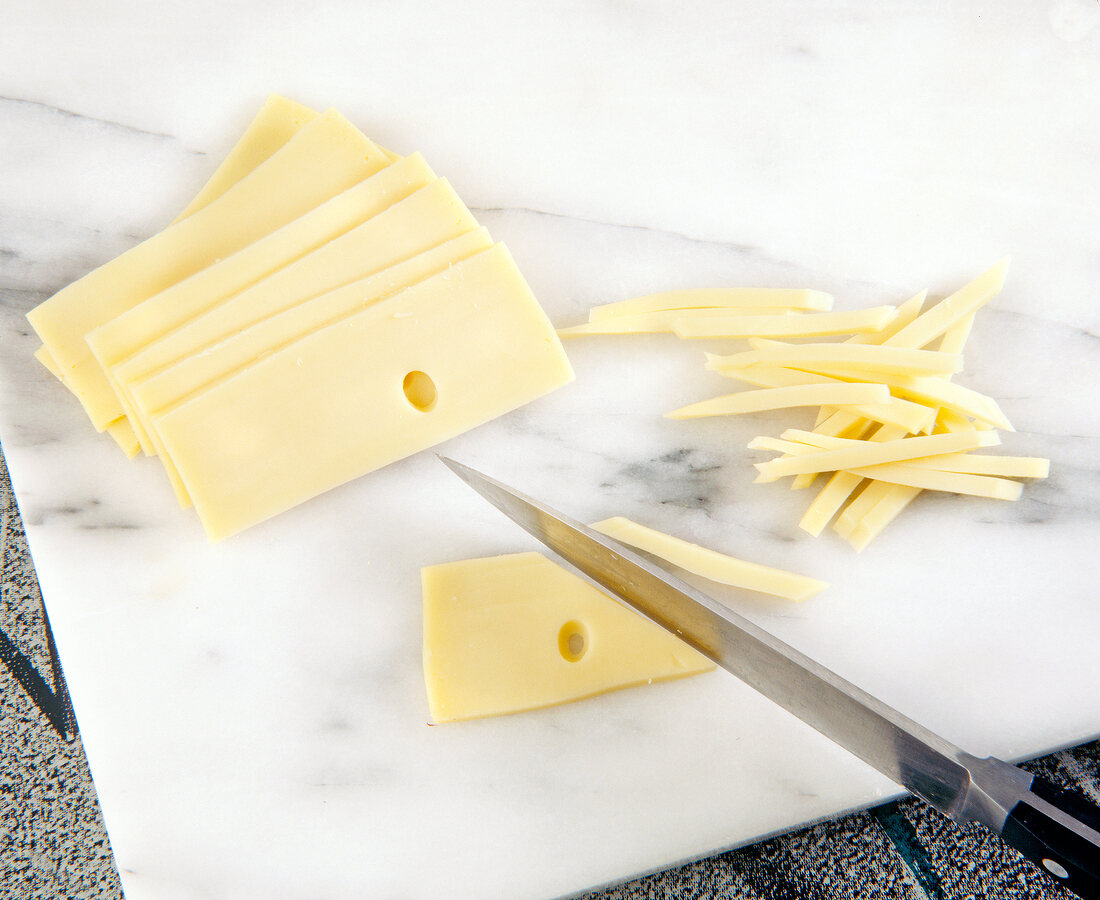 Slices of cheese cut into thin strips with knife on cutting board