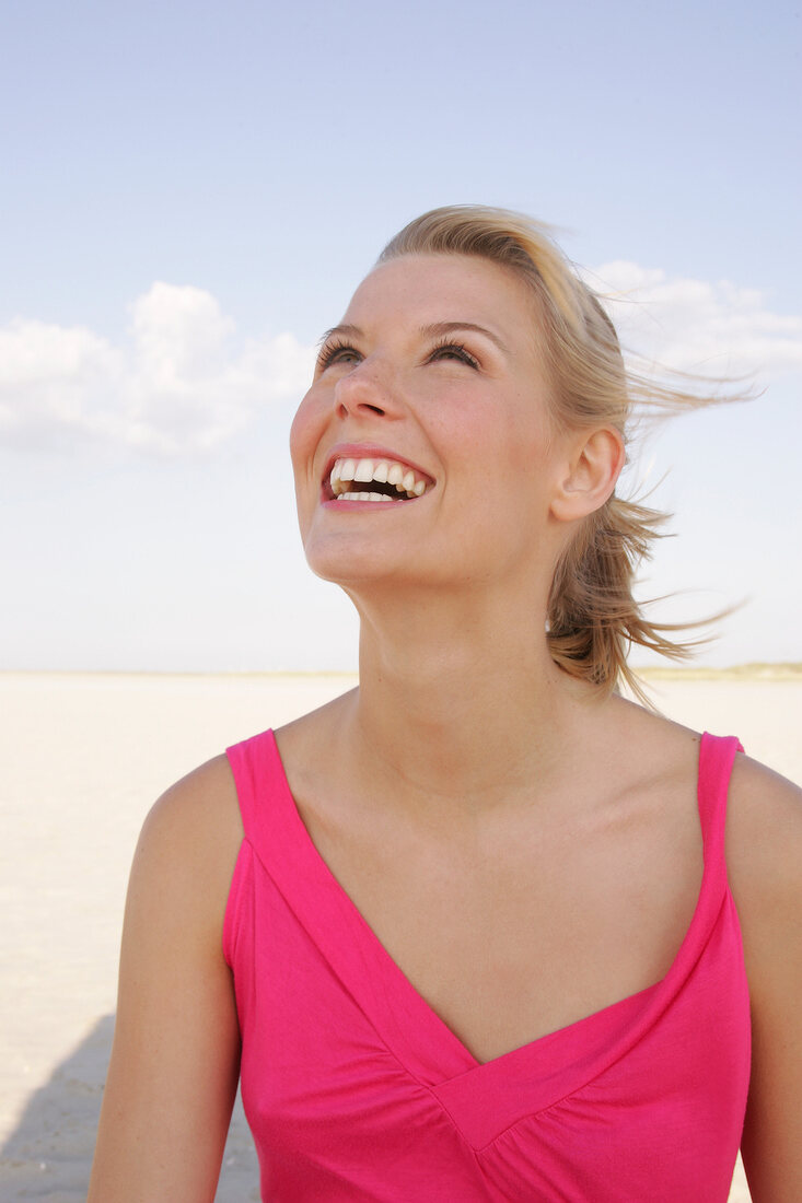 Happy blonde woman in pink dress looking up and smiling on beach