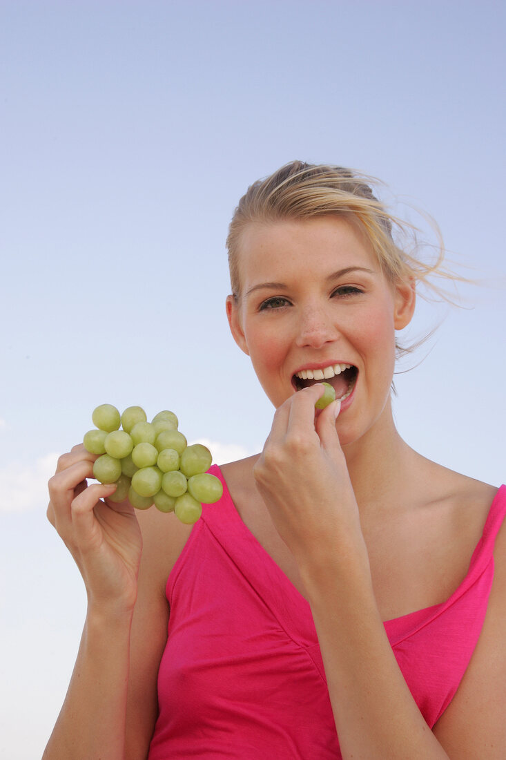 Portrait of blonde woman holding bunch of grapes and eating, smiling