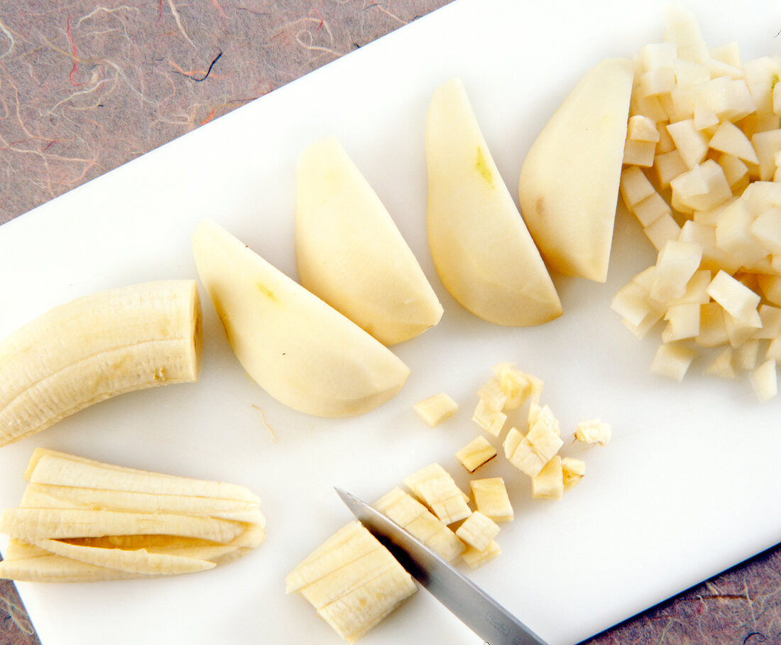 Peeled pears and bananas being chopped in cubes with knife on cutting board