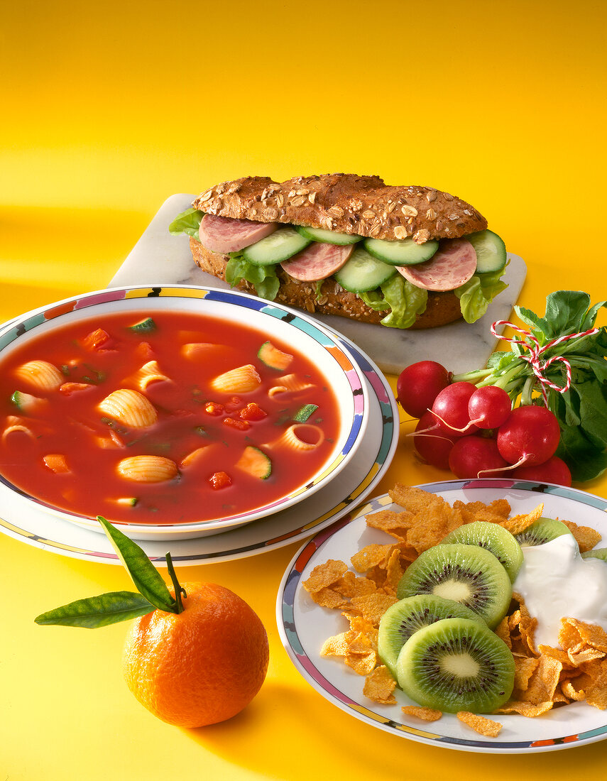 Close-up of vegetable soup, cornflakes with kiwi, sandwich, orange and radishes