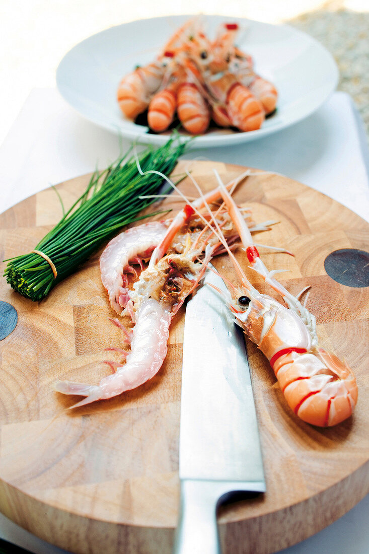 Shrimp, knife and bundle of leek on chopping board, bowl of shrimp in background