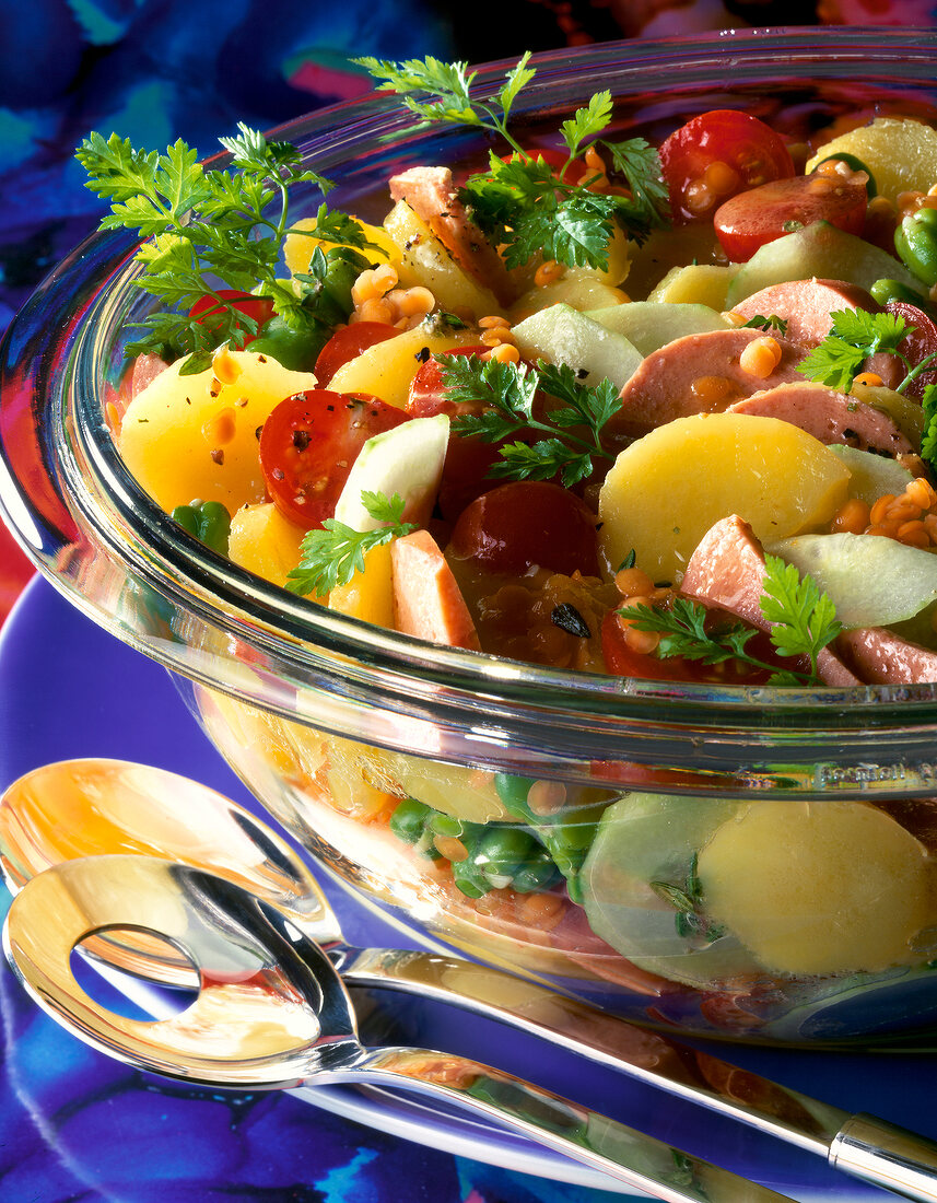 Close-up of potato salad with broad beans, tomatoes, cucumbers and sausage in glass bowl