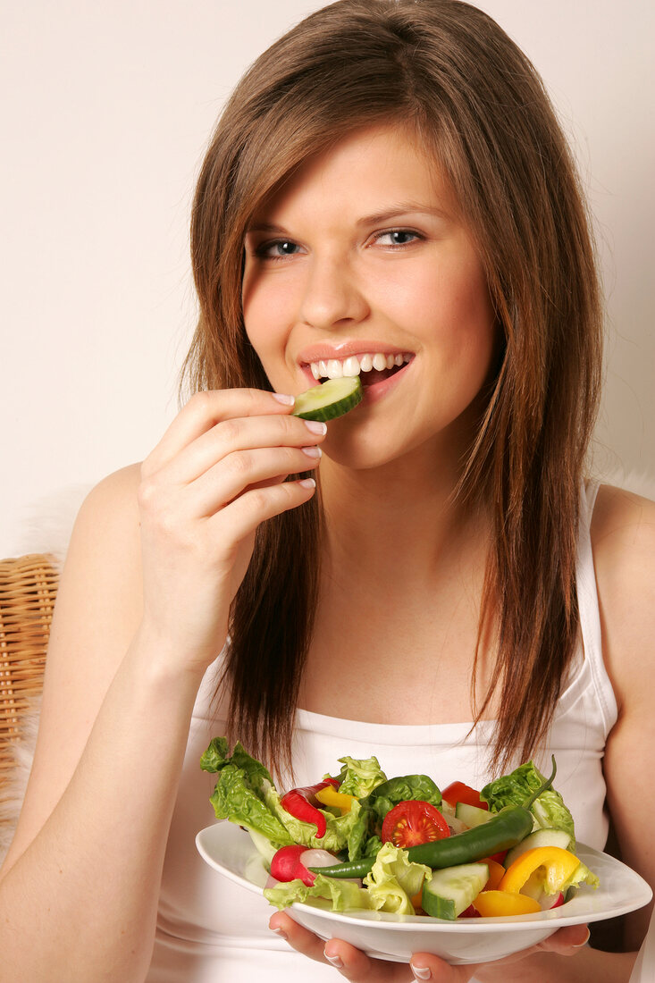 Portrait of woman with long hair holding bowl of salad and eating cucumber, smiling