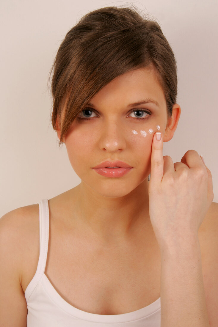 Portrait of pretty woman with brown hair applying cream below her eye with finger