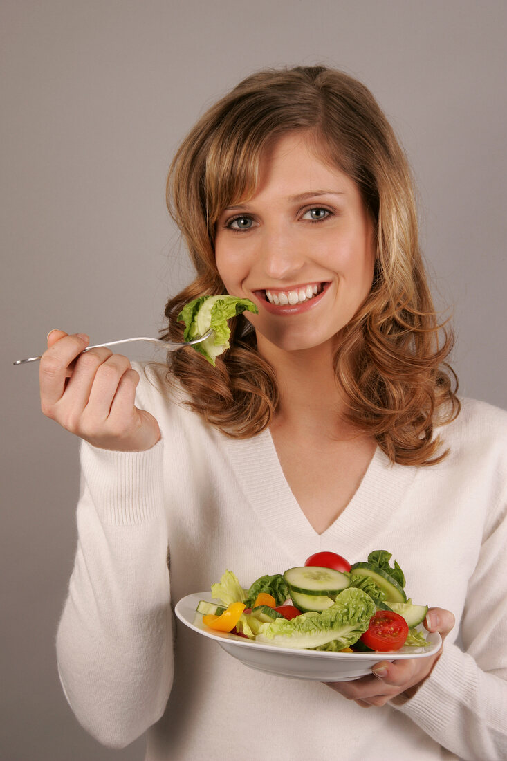 Portrait of beautiful woman eating lettuce with fork while holding salad dish, smiling