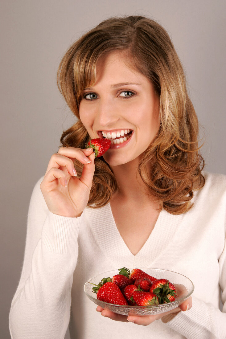 Portrait of young woman biting strawberry while holding plate of strawberries, smiling