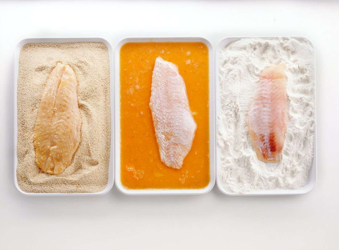 Fish in flour, eggs and bread crumb on three plates, overhead view