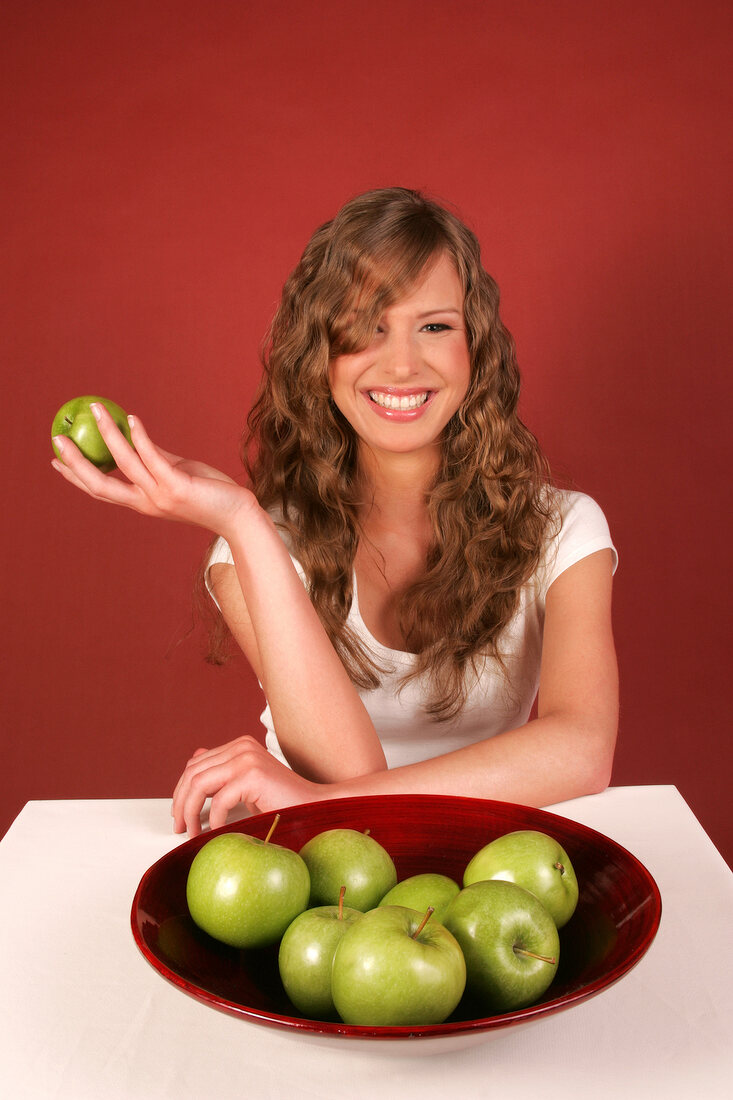Happy woman sitting at table with bowl of green apples and holding apple in hand