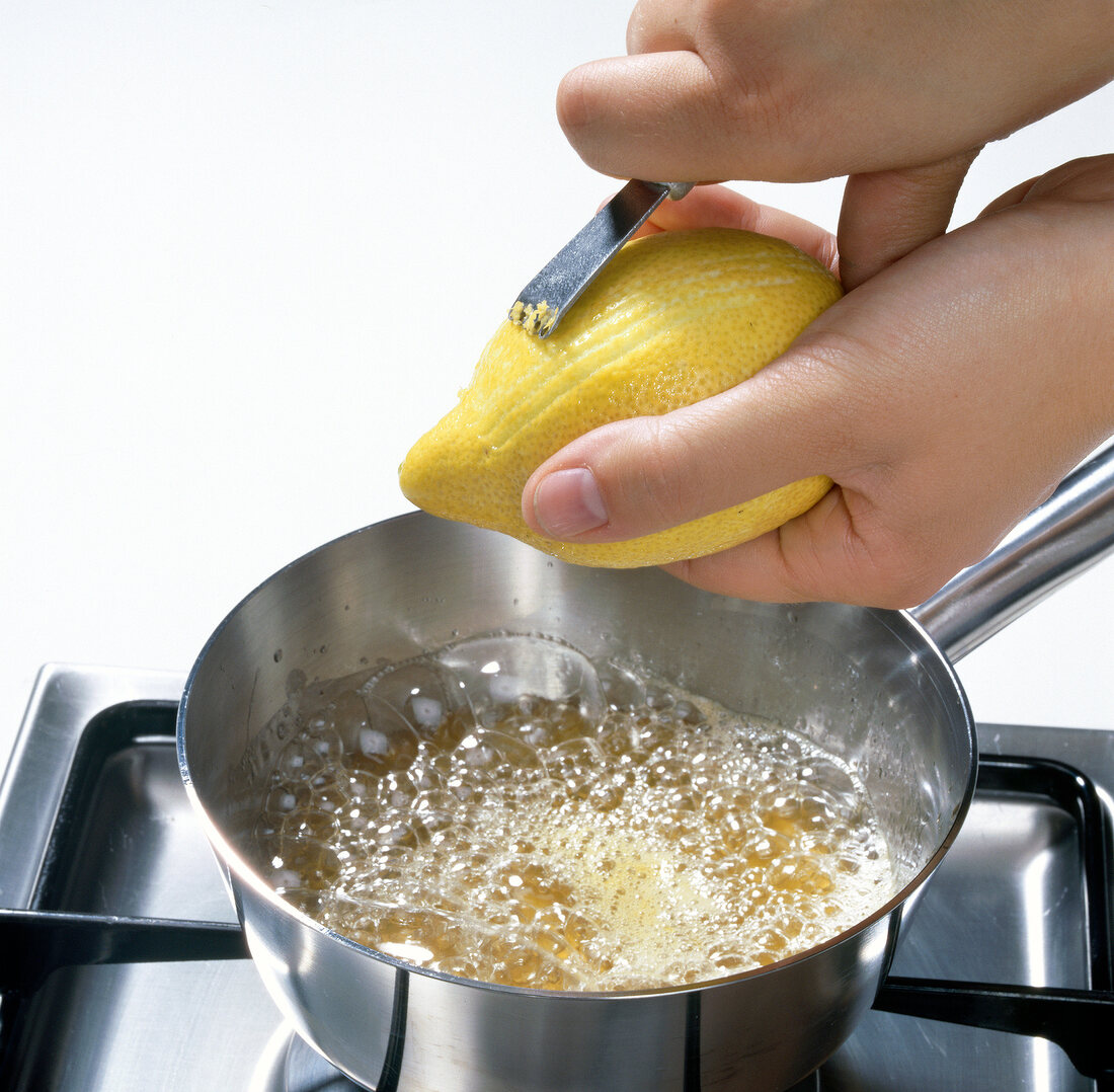 Close-up of hand peeling lemon zest in pot with boiling lemon juice