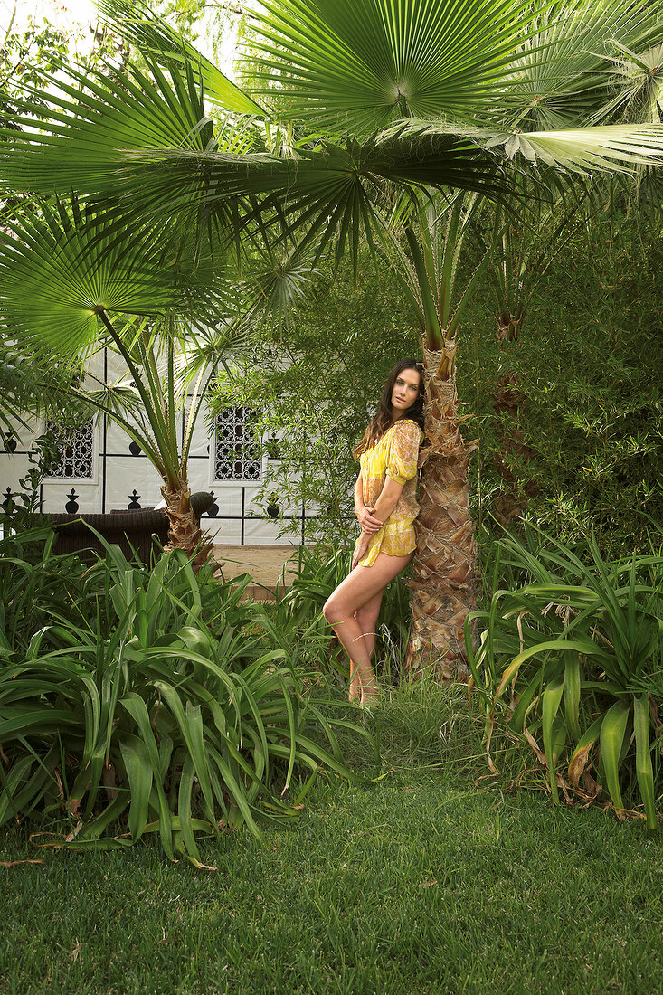 Sexy woman wearing yellow tunic leaning against palm tree in garden